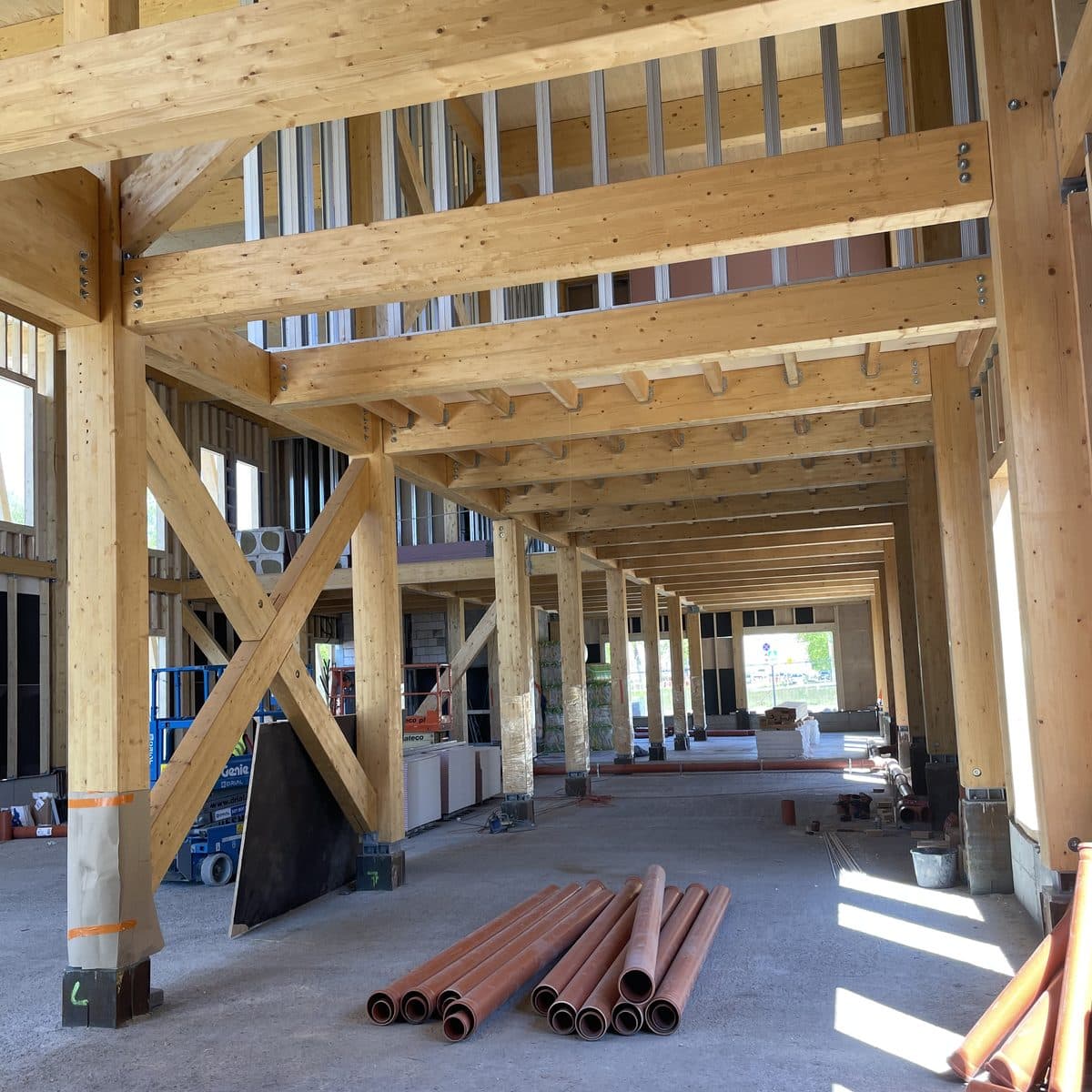 Eine lange Reihe von Stützen, Unterzügen und Kreuzverbänden aus Brettschichtholz im unfertigen Gebäude, auf dem Betonboden liegen Rohre